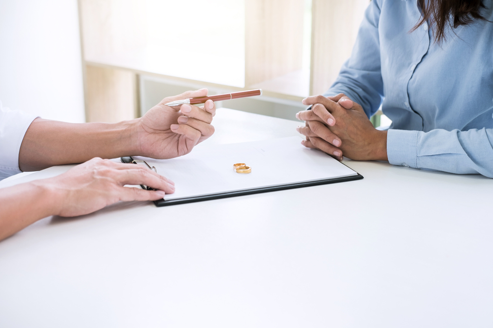 Two people sit across a table. One holds a pen over a clipboard with wedding rings on it. The mood appears serious and focused.