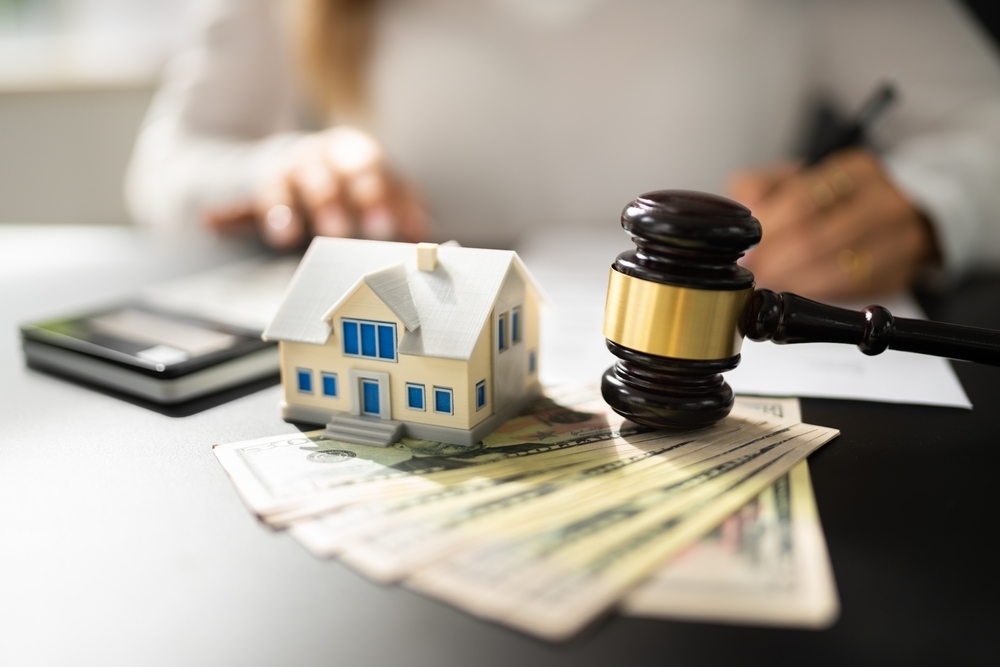 A miniature house sits on a stack of dollar bills next to a judge's gavel. In the background, a person writes, suggesting a legal or financial context.