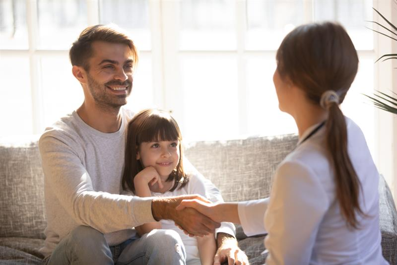 A man and a young girl sit on a sofa, smiling and facing a woman in a white coat. The man is shaking hands with the woman, creating a warm and welcoming atmosphere.