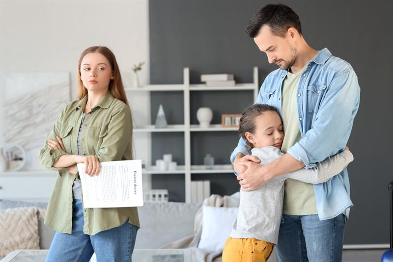 A young girl embraces her father, conveying a sense of comfort, while a woman stands nearby holding divorce papers, looking serious and reflective.