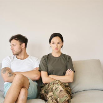 A man and woman sit apart on a sofa with arms crossed, looking in opposite directions, conveying tension. The setting is neutral and minimalistic.