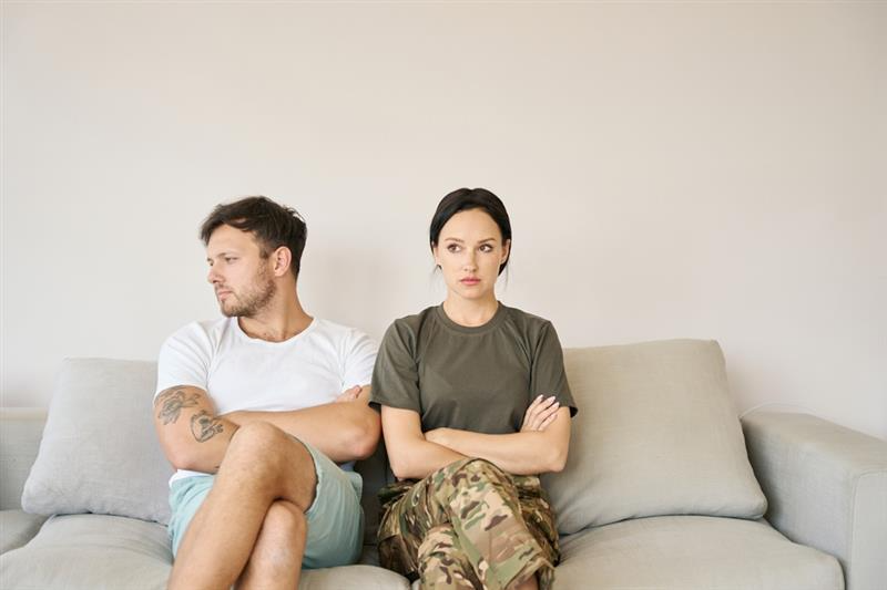 A man and woman sit apart on a sofa with arms crossed, looking in opposite directions, conveying tension. The setting is neutral and minimalistic.