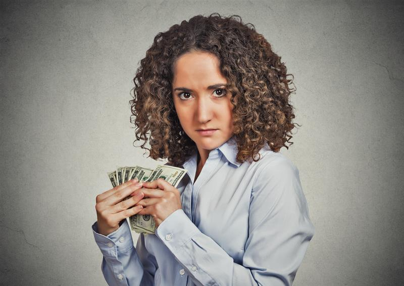 Woman with curly hair holds a stack of cash tightly, wearing a serious expression. Dressed in a white shirt, she stands against a textured gray background.