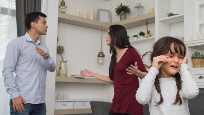 A couple argues in a living room while a young girl in the foreground cries, covering her ears. The scene conveys tension and distress.