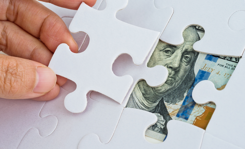 A hand places a white puzzle piece, partially revealing a U.S. dollar bill beneath, with Benjamin Franklin's face visible. It suggests uncovering financial insights.