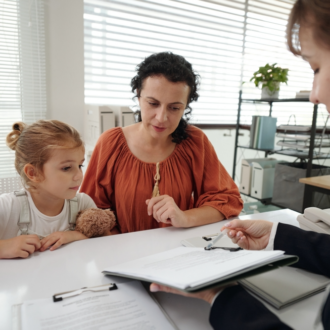 A woman and child sit at a desk with a professional in an office, reviewing documents. The child holds a stuffed animal, creating a serious yet supportive atmosphere.