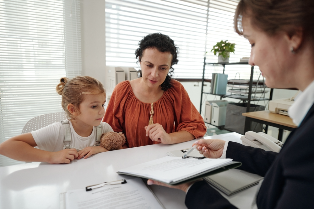 A woman and child sit at a desk with a professional in an office, reviewing documents. The child holds a stuffed animal, creating a serious yet supportive atmosphere.