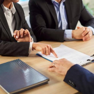 Three professionals in business attire sit at a table reviewing documents. One points at a contract while another holds a pen, suggesting discussions.